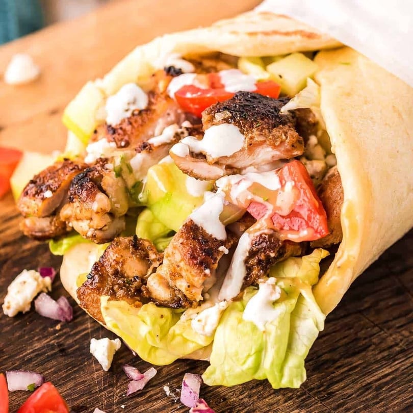 A chicken shawarma wrap with lettuce, tomatoes, and white sauce on a wooden board.
