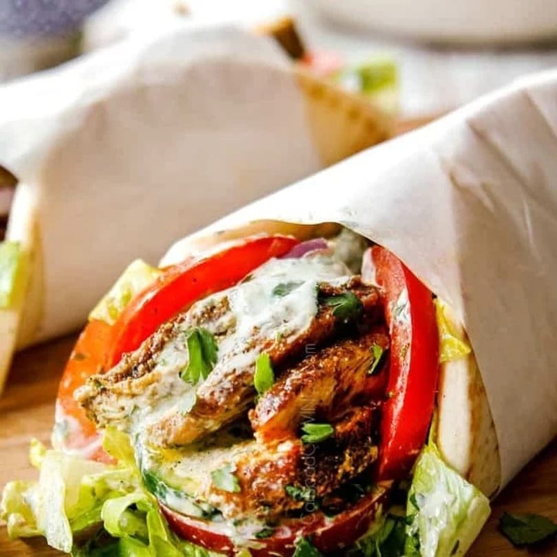 Close-up of a chicken shawarma wrap with tomatoes, lettuce, and white sauce, partially wrapped in paper.